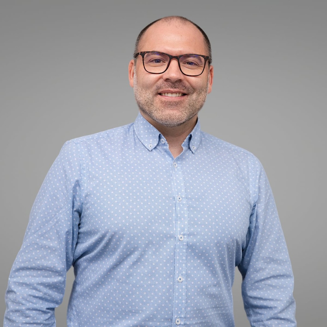 Retrato de estúdio de André Bacêlo e Costa, de camisa azul, sobre fundo cinzento